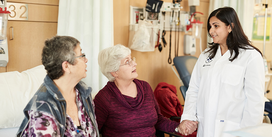 Cathy Chapman and her sister Enid Hollohan sit on a hospital bed and speak with clinical trial coordinator Saara Ali, standing